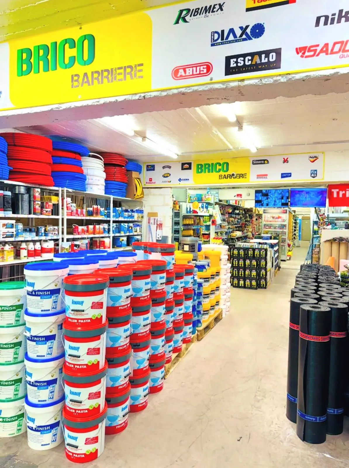 Une allée de Brico Barrière à Saint-Gilles, sous l'enseigne du magasin : pots d'enduit Knauf, rouleaux de roofing, tuyaux et rayons de droguerie.